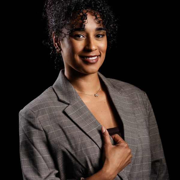 Professional headshot of a woman with curly hair in a grey blazer looking confidently at the camera in a studio setting